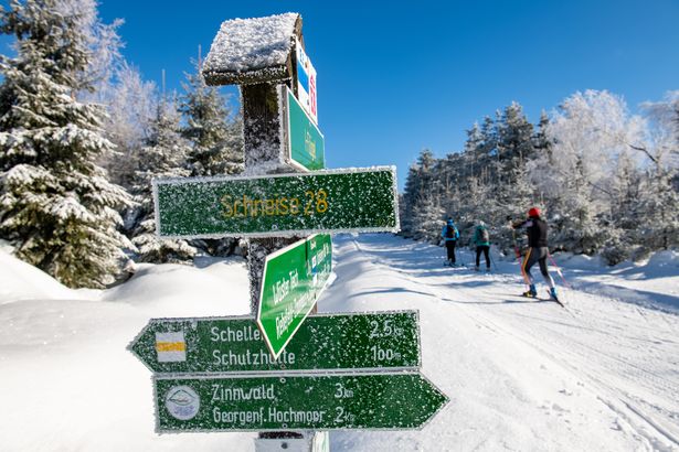 verschneite Langlaufloipe mit Sportlern und Wegweiser Schild im Vordergrund