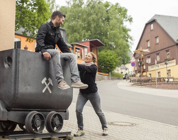 Mann lächelt und sitzt in einem Förderwagen und eine Frau versucht ihn zu schieben. 