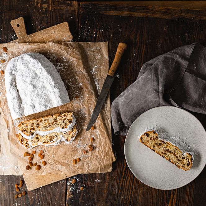 Ein Dresdner Christstollen liegt auf einem Holzbrett. Der Dresdner Stollen wurde mit einem Messer angeschnitten. Auf einem Teller befindet sich eine Scheibe des Dresdner Christstollens. 