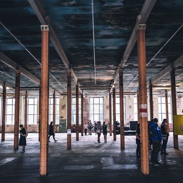 Man sieht ein altes leer stehendes Fabrikgebäude von innen. Die große Halle wird von rostbraunen Pfeilern gestützt. Im Raum verteilt stehen Menschen, die so scheinen, als würden sie sich irgendetwas anschauen.