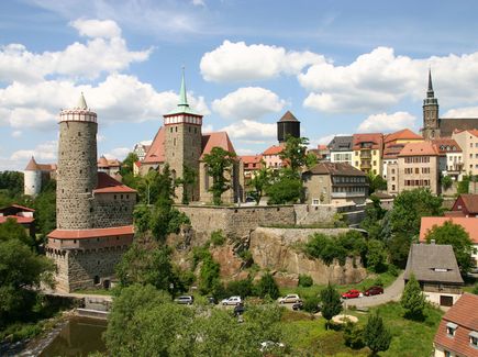 Die Altstadt von Bautzen ist zu sehen mit Türmen und alten Häuser. Ebenso sind Autos, Bäume und Wiesen zu erkennen.