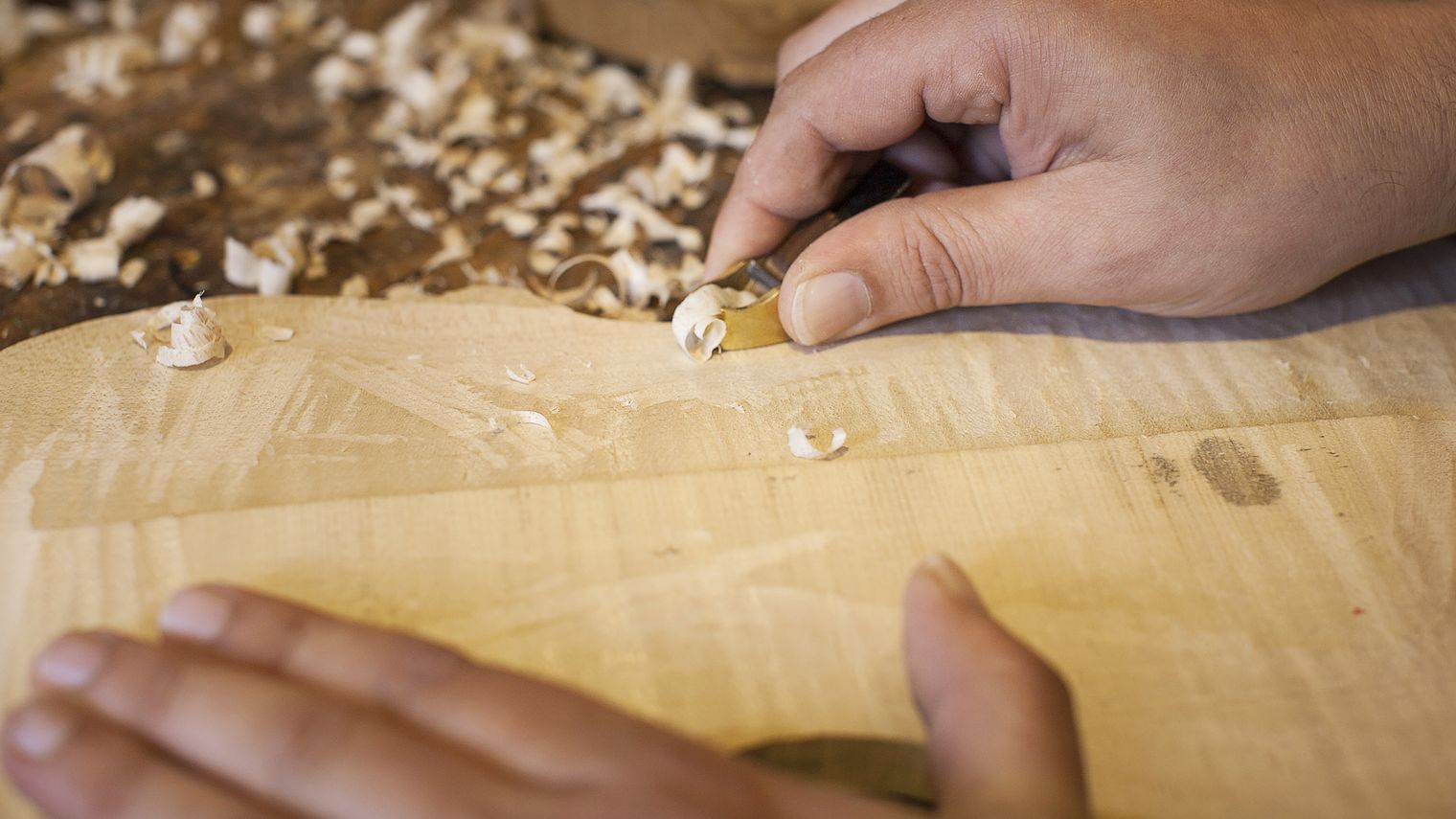 In Handarbeit wird mit einem Werkzeug ein Stück helles Holz bearbeitet.