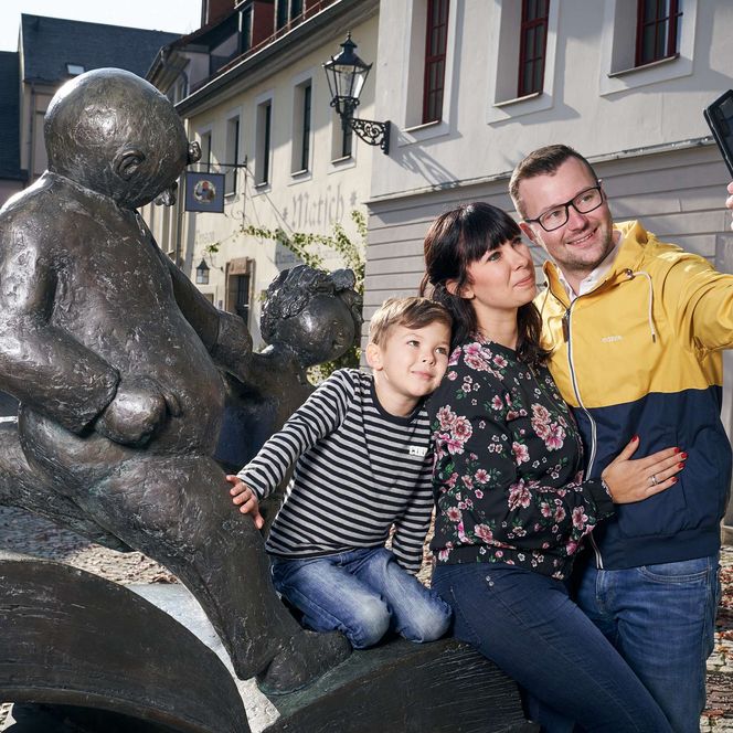 Eine Familie steht an einer Figur in Plauen. Sie machen ein Selfie von ihrem Familienurlaub. 