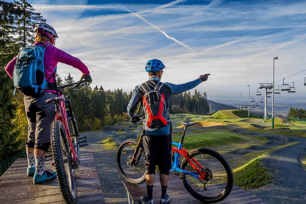 Zwei Mountainbiker stehen am Start eines Trails in der Bikewelt Schöneck. Einer zeigt in die Landschaft des Vogtlandes und zum Sessellift.
