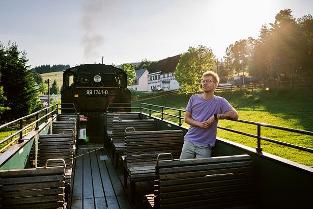 Ein junger Mann fährt im offenen Wagen der Fichtelbergbahn durchs landschaftlich reizvolle Erzgebirge. Er lehnt leicht mit dem Rücken an der Begrenzung des Außenwagens. 