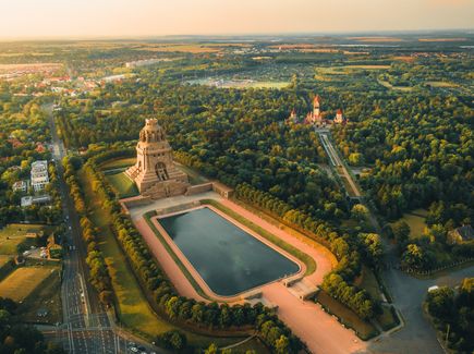 Das Voelkerschlachtdenkmal in Leipzig von oben. Um das Denkmal herum befinden sich Wälder und Häuser. Vor dem Denkmal ist ein kleiner See.