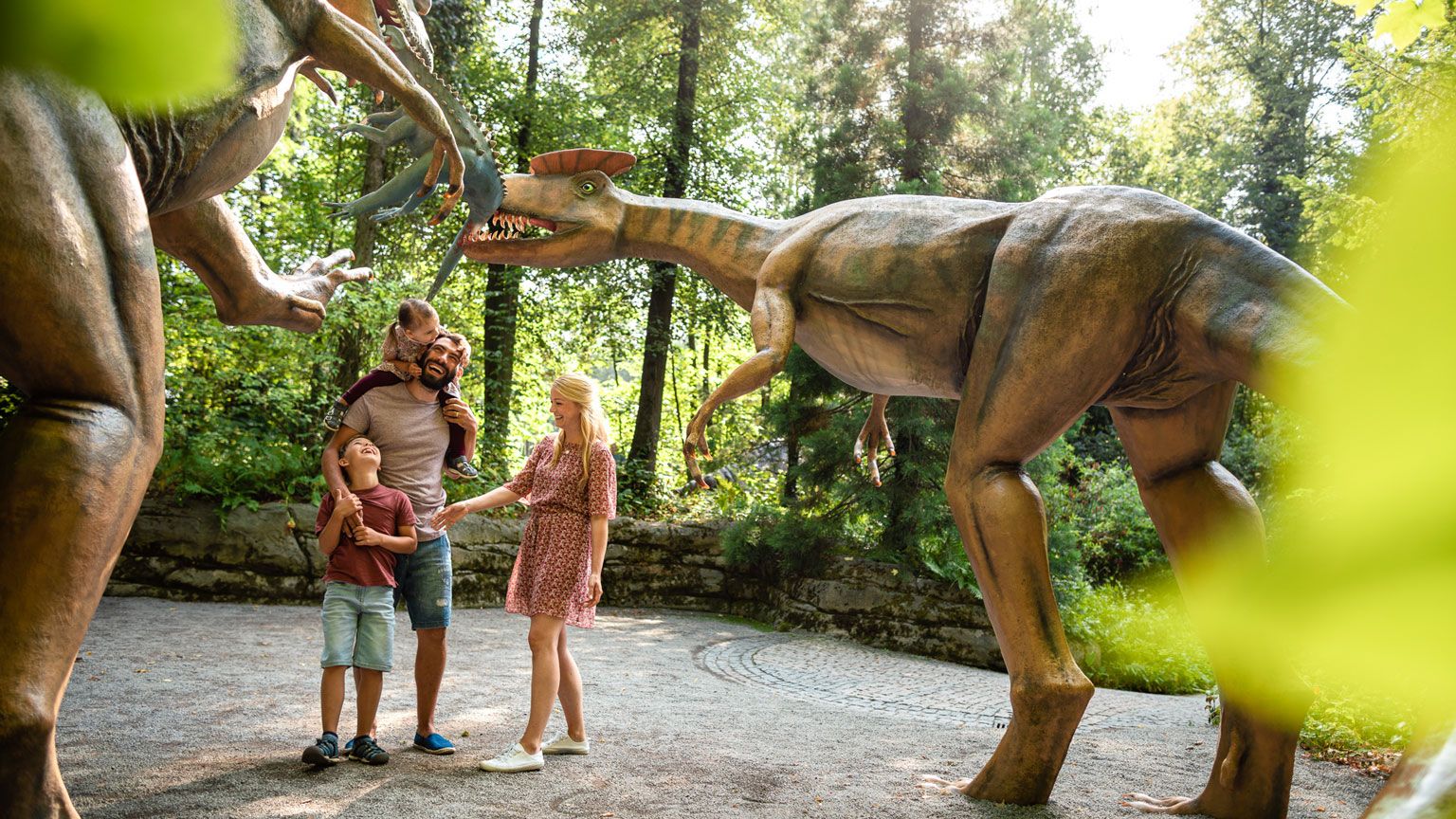Eine Familie bestaunt zwei Dinosaurier in Originalgröße. Sie stehen im Saurierpark.