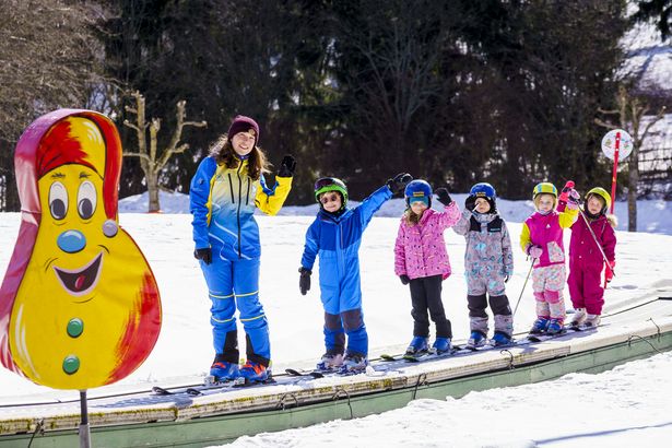 Eine junge Skilehrerin und fünf kleine Skischulkinder stehen auf dem Zauberteppich und winken fröhlich. Daneben sieht man eine bunte Kinderfigur.