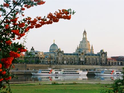 Das Dresdner Elbufer mit der Frauenkirche ist auf dem Bild. Auf der Elbe sind zwei Schiffe, auf der linken Seite des Bildes ein Baum und im Vordergrund eine Wiese. 