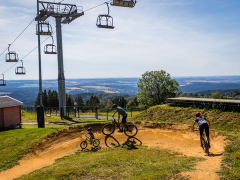 Ein kleines Kind fährt mit seinen Eltern einen Weg in der Bikewelt in Schöneck hinunter. Darüber schweben die Sessellifte und im Hintergrund ist die Landschaft des Vogtlands zu sehen.