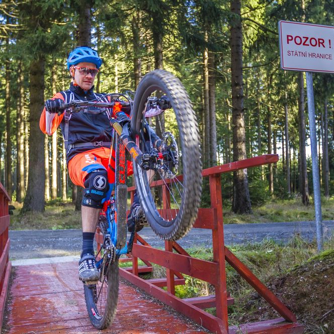Mountainbiker fährt nur auf dem Hinterrad über eine Brücke im Wald. Er ist auf dem MTB-Track Stoneman Miriquidi unterwegs. Er trägt Helm und MTB-Ausrüstung.