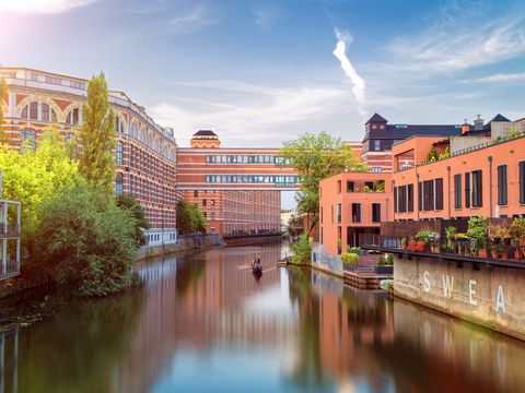 Auf dem Bild ist ein Wasserkanal in Leipzig zu sehen, auf dem ein kleines Boot unterwegs ist. Rechts und links stehen backsteinfarbige Industriegebäude und Bäume, die grünen. Es ist schön Wetter, der Himmel strahlt blau.