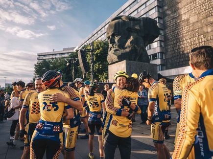 Mehrere Radfahrerinnen und Radfahrer in gelben Jerseys mir Branding Kulturhauptstadt Europas 2025 umarmen sich vor dem Karl-Marx-Monument in der Chemnitzer Innenstadt.