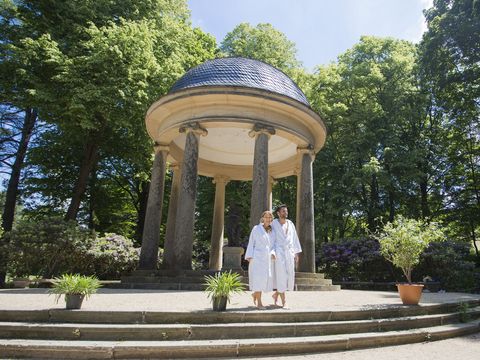 Ein kleiner Tempel steht im Park von Bad Elster. Davor laufen zwei Menschen vorbei.