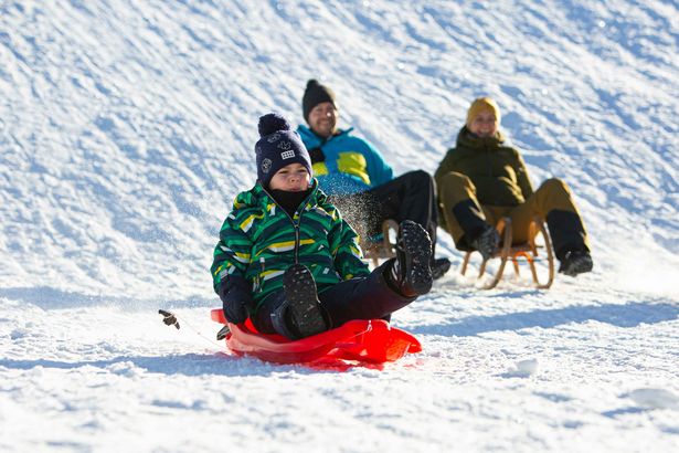 Familie rodelt im Winter einen Berg hinab. Es liegt Schnee und die Sonne scheint.