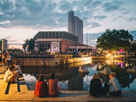 Der Stadthallenpark in Chemnitz abends mit Leute, die an einem Teich sitzen. Im Hintergrund befindet sich die Stadthalle Chemnitz und das hohe Gebäude des Dorint Hotels. Viele Leute befinden sich vor der Stadthalle und es sieht aus als würde ein Fest gefeiert werden.