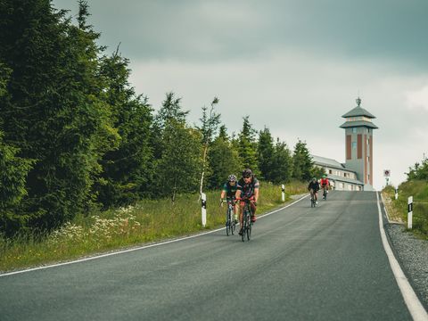 Radfahrer fahren eine Straße herab. Im Hintergrund ist der Turm des Fichtelbergs zu sehen.