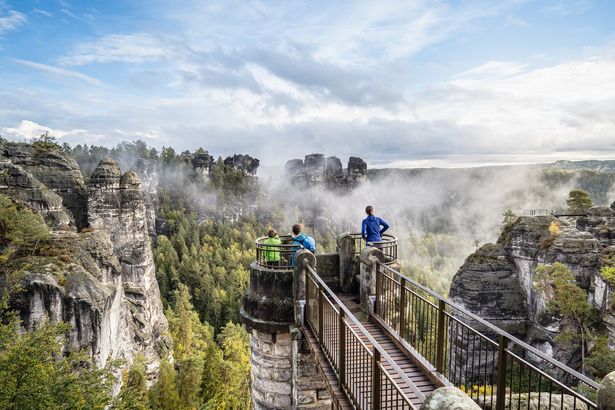 Eine Familie steht auf einer vorgelagerten Aussichtsplattform der Basteibrücke und schaut sich die Sandsteinformationen an. 