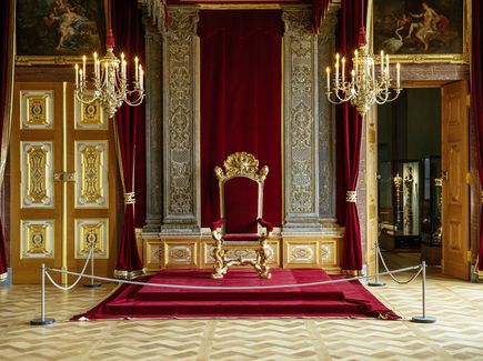 Ein großer, prunkvoller Raum mit einem roten Thron in der Mitte. Von der Decke hängen drei goldene Kronleuchter. 
