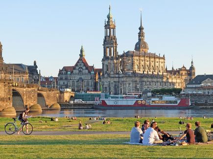 Menschen sitzen am Elbufer in Dresden. Vor ihnen liegt der Elberadweg mit Aussicht auf die Altstadt von Dresden.
